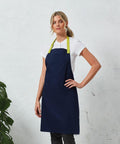 A person wearing a white Premier PR190 shirt and a dark blue apron with green straps, standing against a light textured wall with a plant partially visible on the left.