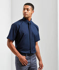 A man wearing a Premier PR202 short-sleeve navy shirt and navy tie stands against a plain white background, looking slightly to the side with one hand in his pocket.