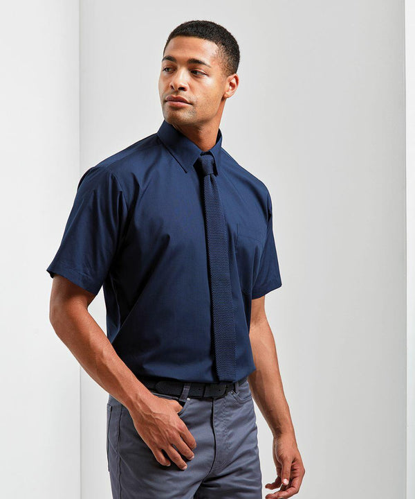 A man wearing a Premier PR202 short-sleeve navy shirt and navy tie stands against a plain white background, looking slightly to the side with one hand in his pocket.