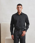A man stands indoors against a neutral background, wearing the Premier Long sleeve Recyclight poplin shirt in black tucked into dark grey trousers with a black belt, facing the camera with a neutral expression and one hand on his waist.