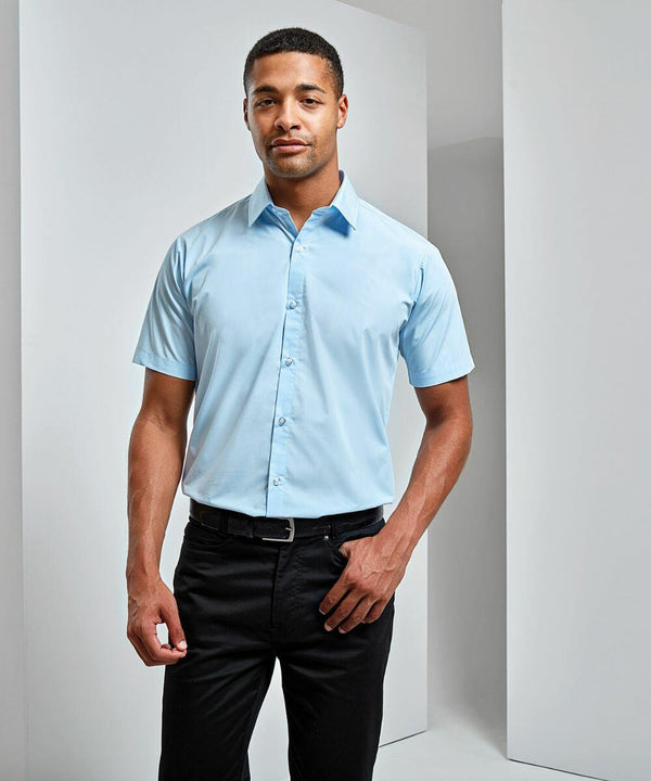 A man stands against a minimalist backdrop, wearing a light blue Premier PR209 short-sleeve button-down shirt and black pants.