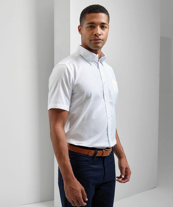 A man wearing a white PR236 short-sleeve button-up shirt by Premier, navy blue pants, and a brown belt stands in a minimalistic setting.