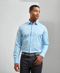 Man in light blue Premier PR244 shirt and dark trousers standing with one hand in his pocket, looking slightly to the side.