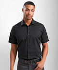 A man wearing a black Premier PR246 short-sleeved button-up shirt and dark gray pants stands against a plain background.