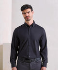 A man with short dark hair and a beard stands against a light-coloured wall, facing forward with a neutral expression. He's wearing the Premier Supreme Oxford long sleeve shirt in black, paired with a black belt and grey trousers.