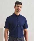 A man with short dark hair and a trimmed beard wears the Premier Supreme Oxford short sleeve shirt in navy, tucked into black trousers, standing against a light-coloured background.