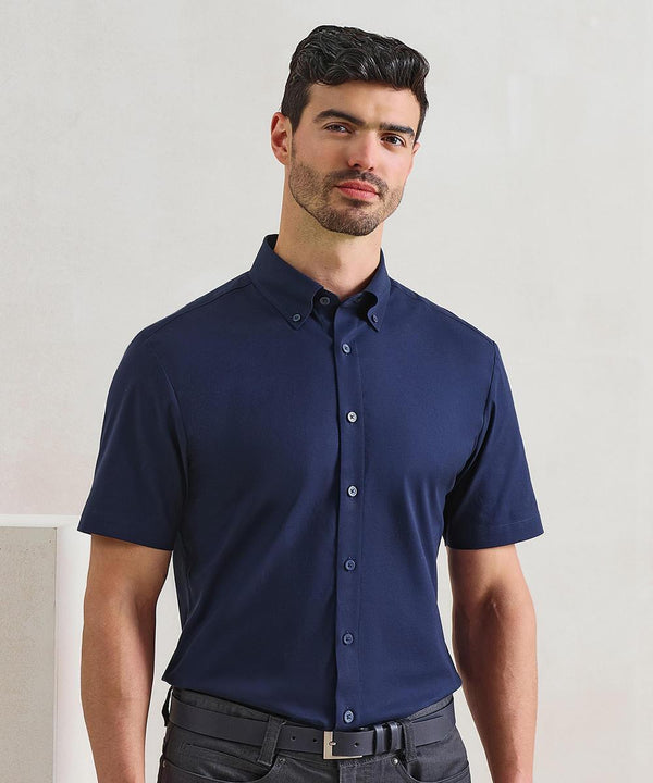 A man with short dark hair and a trimmed beard wears the Premier Supreme Oxford short sleeve shirt in navy, tucked into black trousers, standing against a light-coloured background.