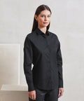 A woman with straight brown hair wears the Premier Women's long sleeve Recyclight poplin shirt in black, standing indoors against a light wall and looking at the camera with a neutral expression.