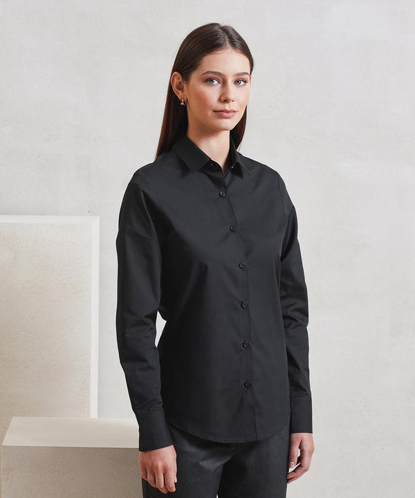 A woman with straight brown hair wears the Premier Women's long sleeve Recyclight poplin shirt in black, standing indoors against a light wall and looking at the camera with a neutral expression.