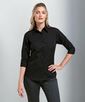 Person standing with one hand on their hip, wearing a black PR305 button-up shirt by Premier with rolled-up sleeves and gray pants, against a plain background.