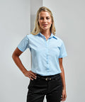 A woman with blonde hair is wearing a light blue PR309 short-sleeve button-up shirt from Premier and black pants, standing against a plain light-colored background.