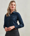 A person with long blond hair is wearing a dark blue Premier PR322 shirt and dark gray pants, standing against a plain light-colored background.
