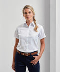 A person stands wearing a white Premier PR336 short-sleeve button-up shirt tucked into dark blue pants, with a brown belt.