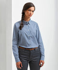 A woman with long braided hair is wearing a Premier PR352 blue checkered shirt and dark gray pants, standing against a plain white background.