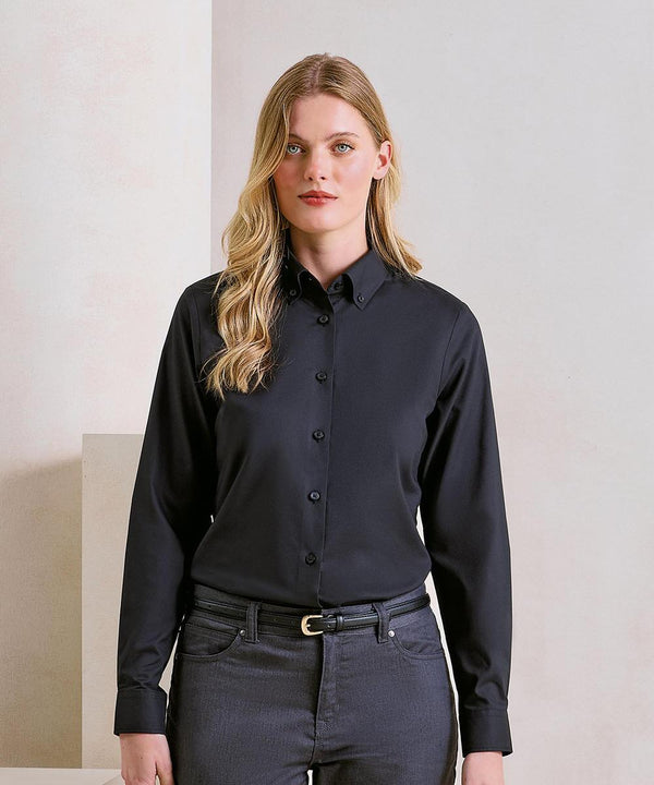 A woman with long blonde hair wears the Premier Women’s Supreme Oxford long-sleeved shirt in black and dark grey trousers, standing against a neutral background and facing the camera with a neutral expression.