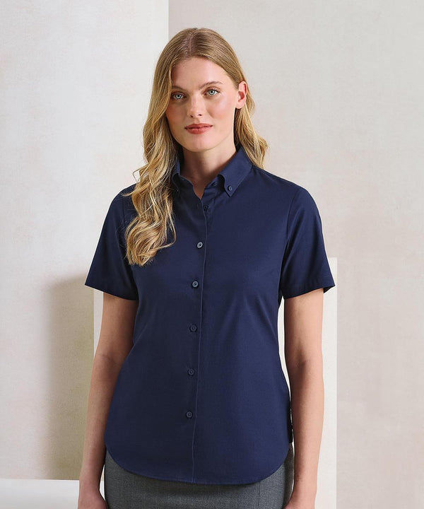 A woman with long blonde hair stands indoors against a neutral background, wearing the Premier Women's Supreme Oxford short sleeve shirt in navy and grey trousers. She faces forward with a neutral expression.