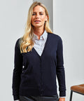 A woman with long blond hair wearing a white shirt and navy blue Premier PR402 cardigan stands indoors, smiling slightly.
