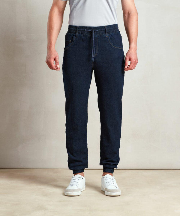 A person standing and wearing a white t-shirt, dark denim jogger pants, and Premier PR556 sneakers, with their hands in their pockets against a neutral background.