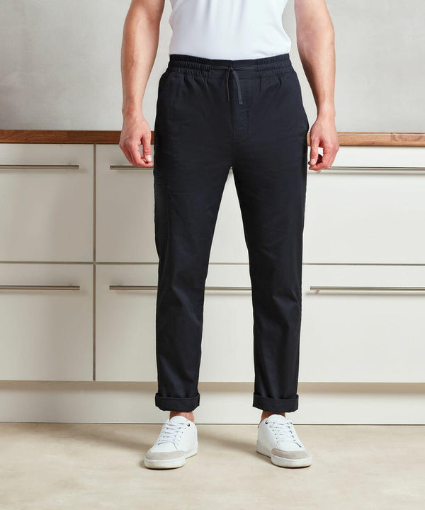 A person stands in a kitchen, wearing a white shirt, Premier PR557 drawstring pants, and white sneakers. The Premier PR557 pants are slightly rolled up at the ankles.