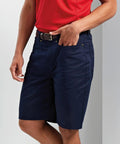 A person is wearing a red Premier PR562 shirt, navy blue shorts with pockets, and a black belt. They are standing against a light background.