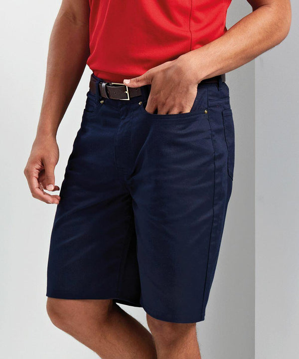 A person is wearing a red Premier PR562 shirt, navy blue shorts with pockets, and a black belt. They are standing against a light background.