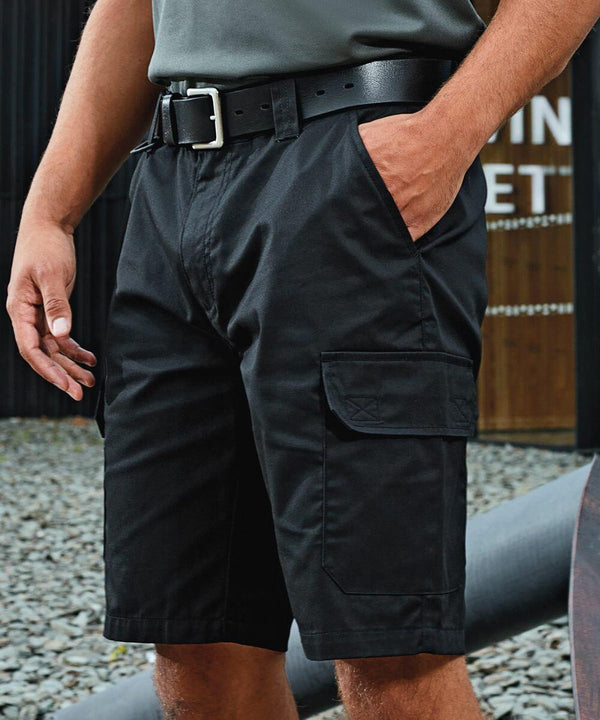 A person wearing black Premier PR564 cargo shorts with a belt stands with hands in pockets. The background features gravel and a building with partially visible signage.