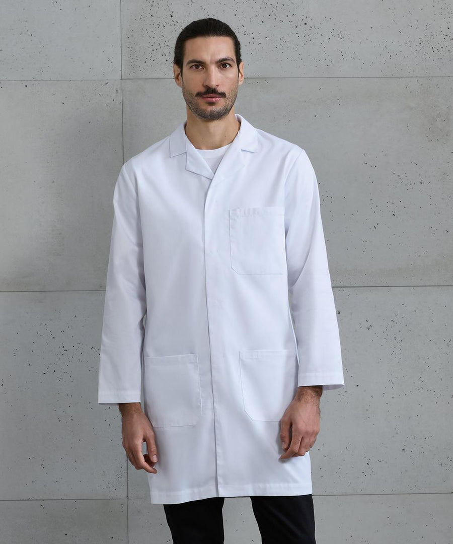 A man with dark hair and a beard stands against a grey concrete wall, wearing Premier's Essential 3-pocket work coat over a white shirt and black trousers.