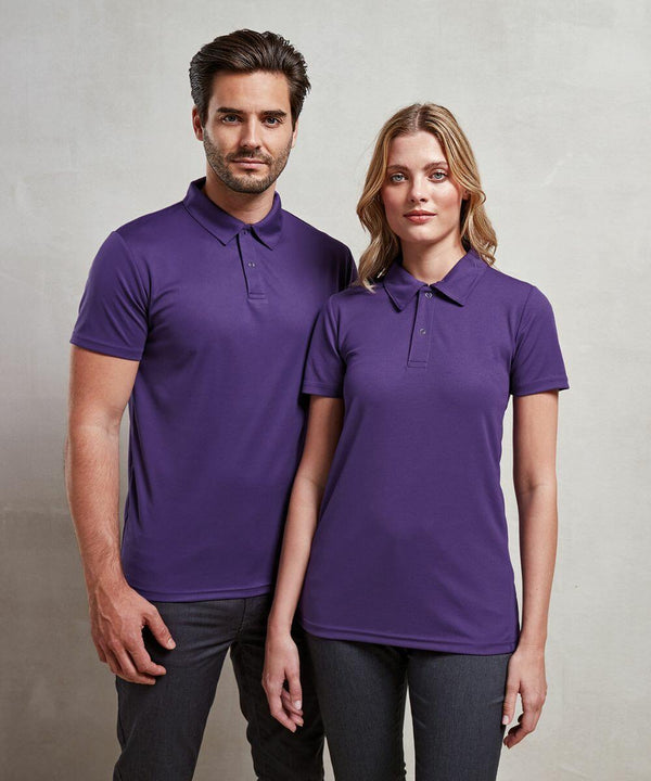A man and a woman stand side by side wearing matching purple Premier PR612 polo shirts and dark pants, posing against a neutral background.