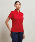 Person wearing a red Premier PR616 short-sleeve polo shirt and dark pants, standing with one hand on hip against a plain backdrop.