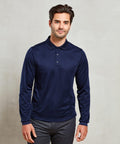 A man with dark hair and a beard stands against a plain background wearing a long-sleeved navy blue Premier PR617 polo shirt and gray pants.