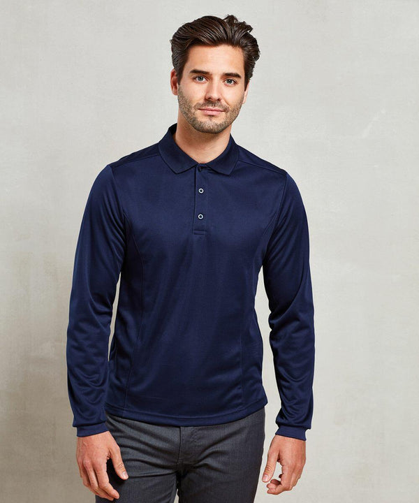 A man with dark hair and a beard stands against a plain background wearing a long-sleeved navy blue Premier PR617 polo shirt and gray pants.
