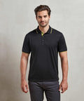 A man with short hair and a beard wears a black Premier PR618 polo shirt with yellow accents and dark pants, standing against a plain light-colored background.