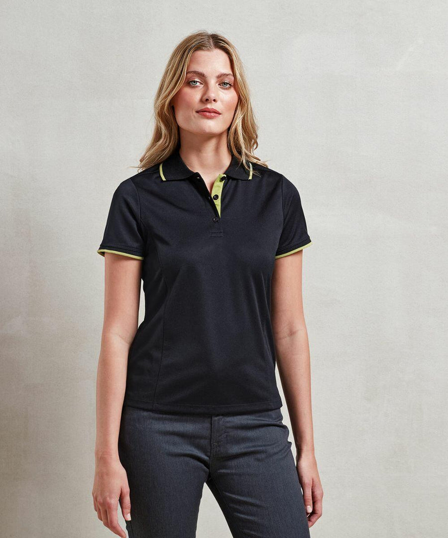Woman with long wavy hair wearing a black Premier PR619 polo shirt and dark gray pants standing against a plain light gray background.