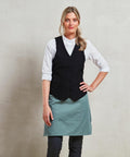 A person stands against a plain background wearing a white shirt, black vest, and green Premier PR623 apron, looking forward with one hand on their hip.