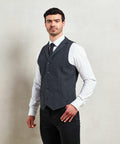 A man stands against a plain background wearing a grey Premier PR625 waistcoat over a white shirt and black tie, with his hand resting on his hip.