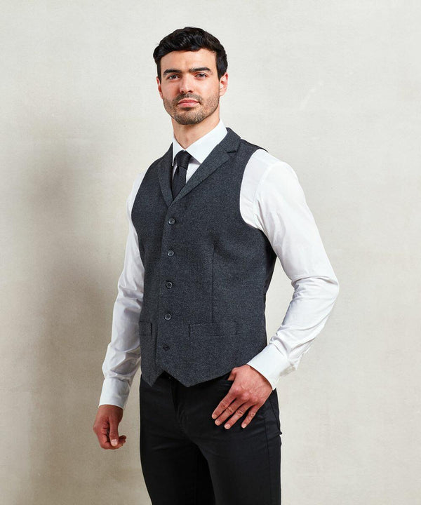 A man stands against a plain background wearing a grey Premier PR625 waistcoat over a white shirt and black tie, with his hand resting on his hip.
