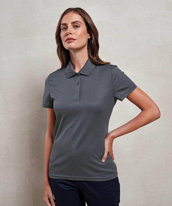 A woman wearing a dark gray Premier PR633 polo shirt stands with one hand on her hip against a plain background.