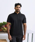 A man with short, curly dark hair and a beard stands indoors wearing the Premier Essential Coolchecker short sleeve chefs' shirt in black and dark trousers. Behind him, vegetables and tinned goods sit on a metal table.