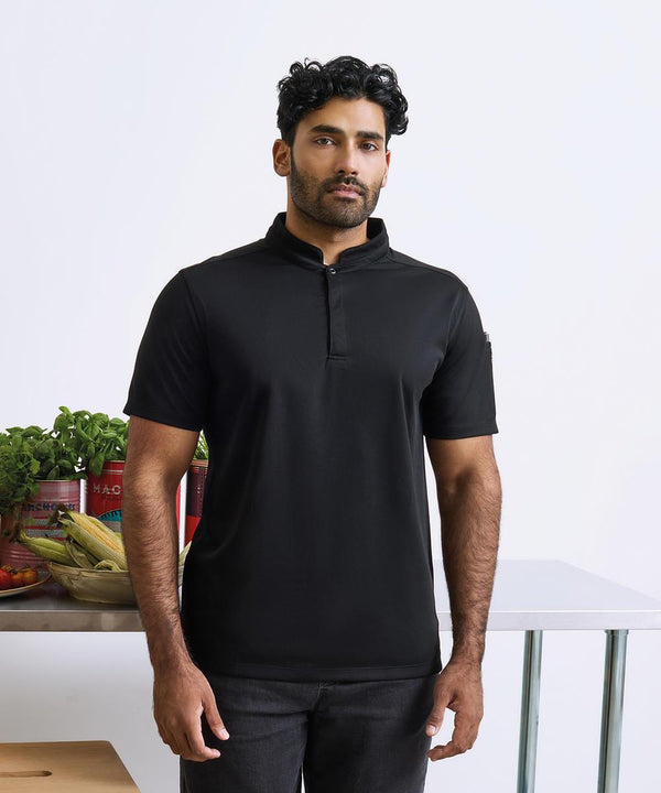 A man with short, curly dark hair and a beard stands indoors wearing the Premier Essential Coolchecker short sleeve chefs' shirt in black and dark trousers. Behind him, vegetables and tinned goods sit on a metal table.