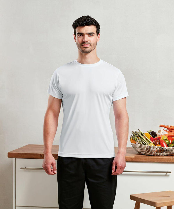 A man wearing a plain white Premier PR649 t-shirt and black pants stands in a kitchen next to a wooden counter with a basket of assorted vegetables.