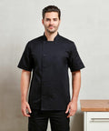 A person in a black short-sleeved Premier PR656 chef coat stands in a kitchen setting with a light gray wall and a wooden countertop.