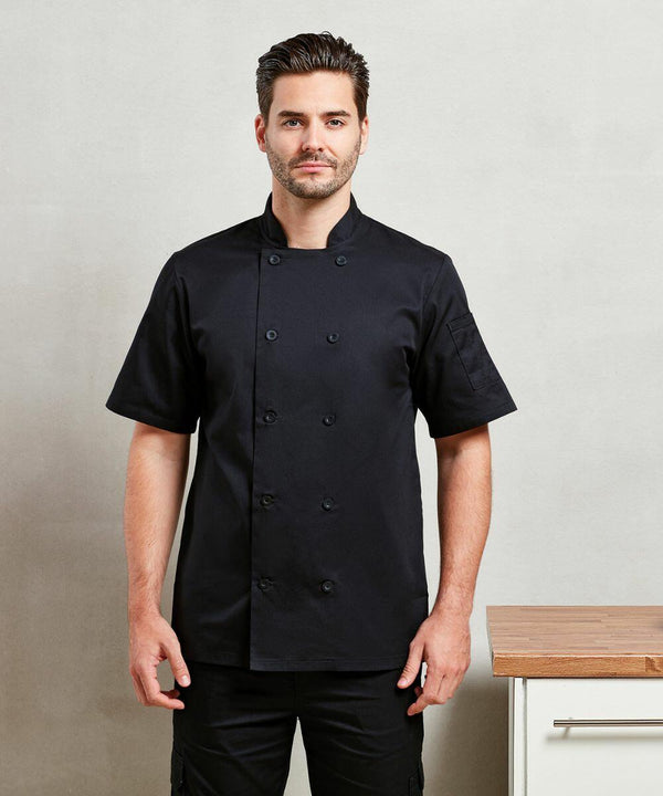 A person in a black short-sleeved Premier PR656 chef coat stands in a kitchen setting with a light gray wall and a wooden countertop.