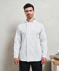 A person stands wearing a white double-breasted chef's jacket, Premier PR657, facing the camera, with a neutral expression. A countertop with stacked dishes is partially visible on the right.