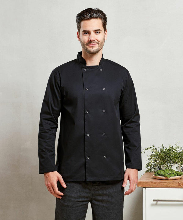 A man in a black double-breasted Premier PR665 chef's jacket stands in front of a light-colored wall, next to a wooden counter holding a green plant.