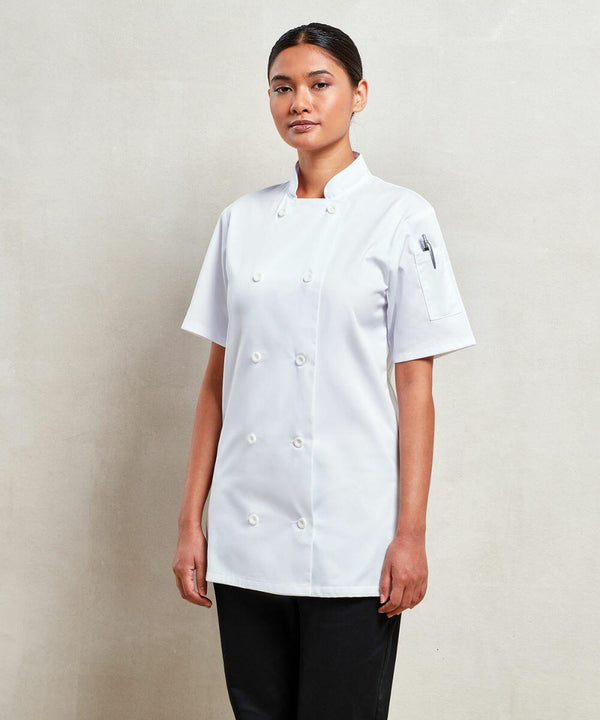 A person stands against a plain background wearing a double-breasted, short-sleeved white Premier PR670 chef's jacket and black pants.