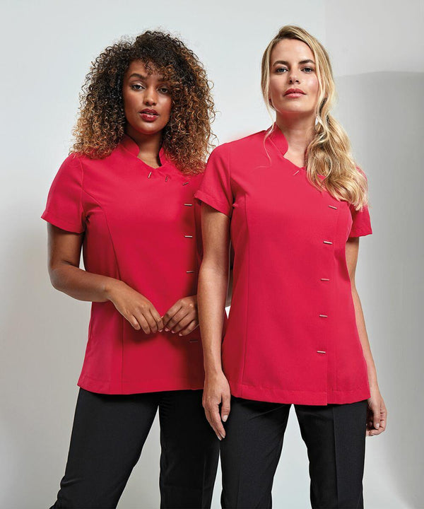 Two women standing side by side wearing matching Premier PR682 red button-up shirts and black pants. The woman on the left has curly hair, and the woman on the right has straight hair tied back.
