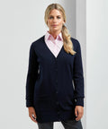 A woman stands against a plain white background, wearing a light pink collared shirt under a buttoned-up navy blue cardigan. She has blonde hair tied in a side ponytail and is looking straight ahead, showcasing the Premier PR698.