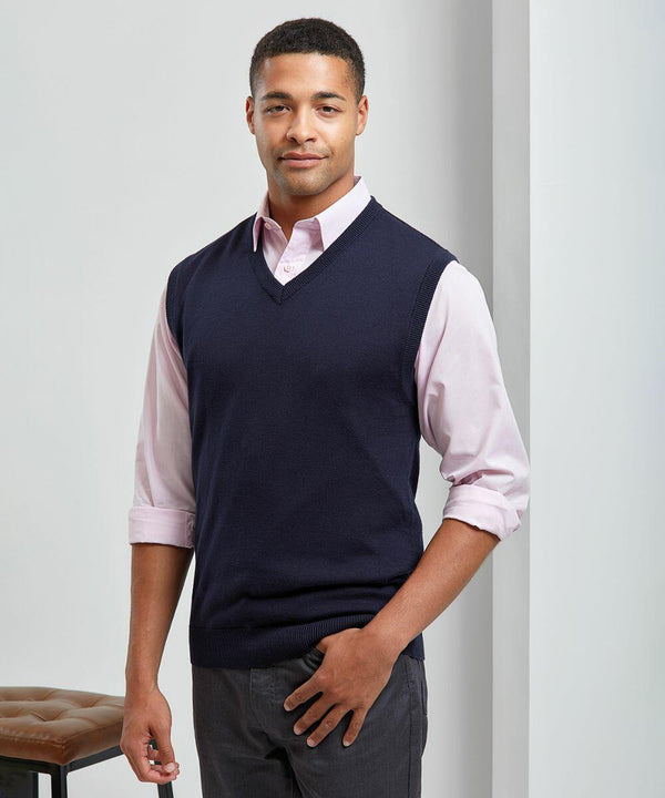 A man wearing a navy blue Premier PR699 sweater vest over a light pink shirt with rolled-up sleeves stands indoors, posing and looking at the camera.