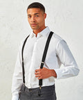 A man wearing a white Premier PR701 dress shirt, grey pants, and black suspenders stands against a white textured background.