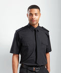 A man in a black Premier PR710 short-sleeve uniform shirt with two chest pockets and a black tie stands against a plain background.